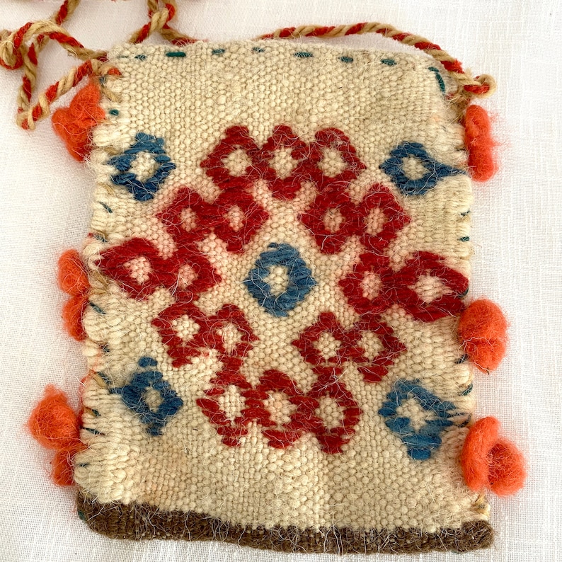 Pu&ograve; includere: Una piccola borsa intrecciata di colore crema con un motivo geometrico in rosso, blu e marrone. La borsa ha una chiusura a coulisse ed &egrave; decorata con pompon rossi.