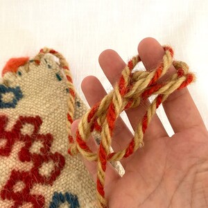 Pu&ograve; includere: Primo piano di un arazzo tessuto beige con ricami rossi e blu. Un cordone spesso con un filo di lana beige e rosso &egrave; annodato.