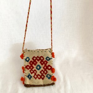 Pu&ograve; includere: Una piccola borsa intrecciata con un motivo geometrico in rosso, blu e bianco. La borsa ha una lunga tracolla e pompon arancioni intorno ai bordi.