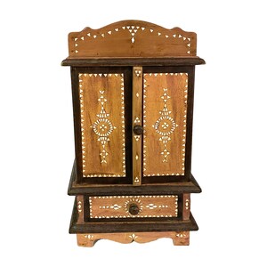 May include: A small wooden cabinet with two doors and a drawer. The cabinet is decorated with intricate geometric patterns made from inlaid bone or shell. The cabinet is brown and has a light wood finish.