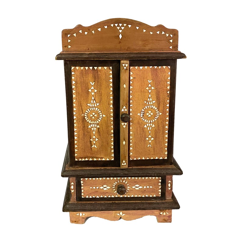 May include: A small wooden cabinet with two doors and a drawer. The cabinet is decorated with intricate geometric patterns made from inlaid bone or shell. The cabinet is brown and has a light wood finish.