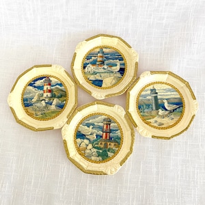 Könnte beinhalten: Set aus vier weißen Keramik-Untersetzern mit maritimem Thema. Jeder Untersetzer zeigt einen Leuchtturm, Möwen und eine blaue und weiße Meereslandschaft. Die Untersetzer haben einen Seil-Rand.