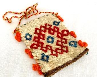 May include: A small, white woven bag with a red and blue geometric pattern. The bag has a brown leather trim and a long, brown cord strap. The bag is decorated with orange pom-poms.
