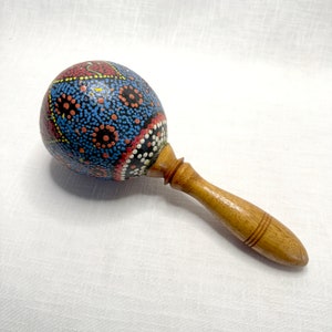 Könnte beinhalten: Eine Holzmaraca mit einem runden, bemalten Kopf. Der Kopf ist mit einem blauen, roten und schwarzen Punktmuster verziert. Der Griff ist aus Naturholz.