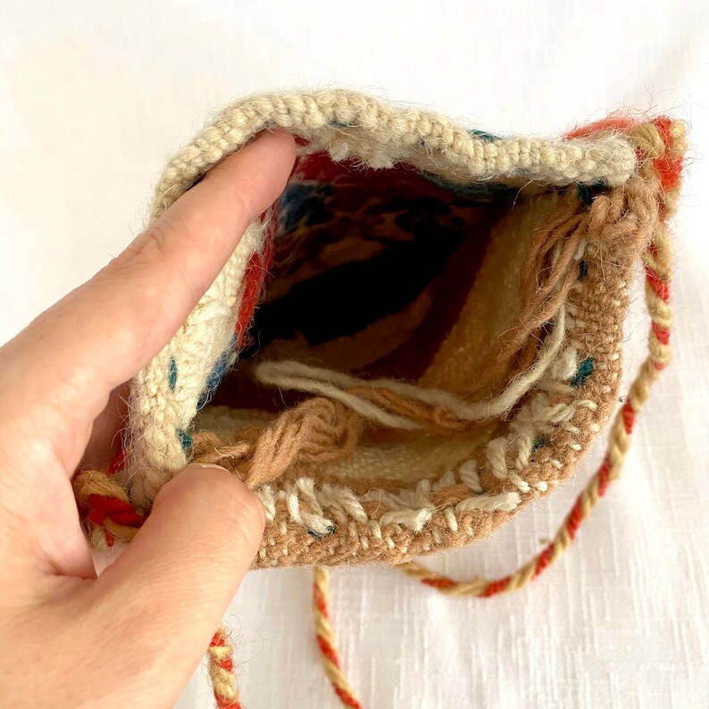 Pu&ograve; includere: Una piccola borsa intrecciata con chiusura a coulisse. La borsa &egrave; realizzata in tessuto beige con accenti rossi, blu e bianchi. La borsa ha una lunga tracolla intrecciata.