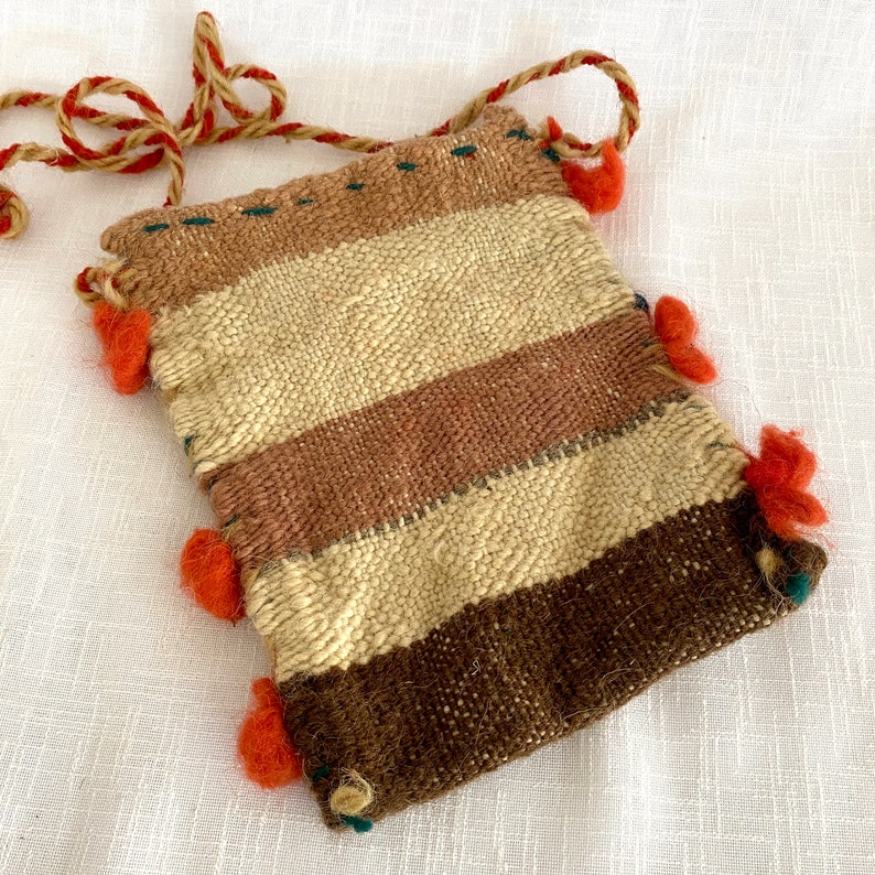 Pu&ograve; includere: Una piccola borsa intrecciata con un motivo a righe marroni, beige e arancioni. La borsa ha una chiusura a coulisse ed &egrave; decorata con pompon arancioni.