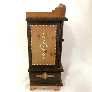 May include: A small wooden cabinet with a dark brown finish and intricate white inlay designs. The cabinet has a single door and a shelf inside. The cabinet is decorated with geometric patterns and a floral motif.