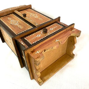 May include: A small wooden cabinet with a hinged door and a drawer. The cabinet is decorated with intricate inlaid patterns of wood and bone. The cabinet is made of light brown wood with dark brown accents.