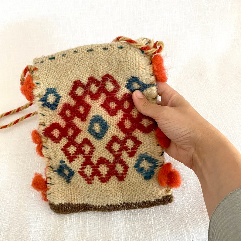 Pu&ograve; includere: Una piccola borsa intrecciata con un motivo geometrico in rosso, blu e bianco. La borsa ha un bordo in pelle marrone e una frangia di filo rosso.