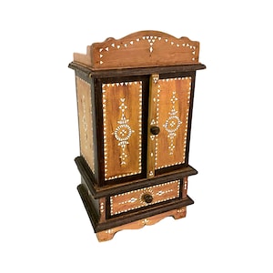 May include: A small wooden cabinet with a decorative inlaid design. The cabinet has two doors and a drawer, all with intricate patterns. The cabinet is painted in shades of brown and has a slightly arched top.