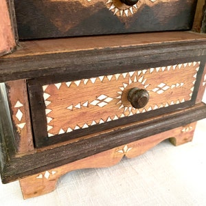 May include: A small wooden box with three drawers. The drawers are decorated with white geometric patterns and have wooden knobs. The box is made of dark brown wood with a light brown finish.