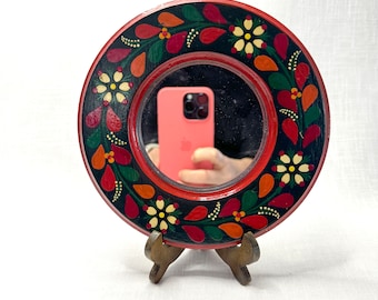May include: A red and black round wooden mirror with a floral design. The mirror has a stand and is reflecting a smartphone.