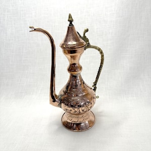 May include: A copper teapot with an ornate design and a brass handle. The teapot has a long spout and a rounded body.