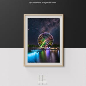 Könnte beinhalten: Ein gerahmter Druck mit einem hell beleuchteten Riesenrad bei Nacht, das sich im Wasser spiegelt. Das Rad ist mit einem Regenbogen von Farben vor einem Sternenhimmel beleuchtet. Das Bild ist in einem Holzrahmen.
