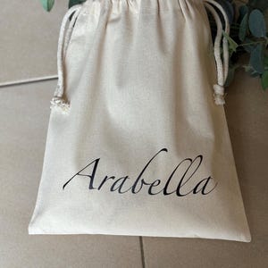 May include: A cream-coloured drawstring bag with the name "Arabella" printed in black script. The bag is made of a soft, natural fabric and has a simple, elegant design. The drawstring is a light cream colour.