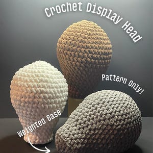 May include: Three crochet display heads in white, tan, and gray. The white head has a weighted base. The tan head is in the center, and the gray head is on the right. The text "Crochet Display Head" and "Pattern Only!" are visible.