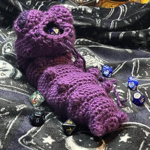 May include: A handmade, purple crochet dice bag with a drawstring closure. The bag is textured and has a unique shape. Several multi-sided dice in various colors are scattered around the bag, suggesting a tabletop gaming theme.