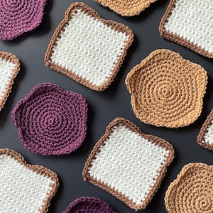 May include: Crocheted coasters in various shapes and colors. Some are shaped like slices of bread with white centers and brown edges. Others are round or petal-shaped in shades of brown and purple. These handmade coasters are perfect for protecting surfaces.