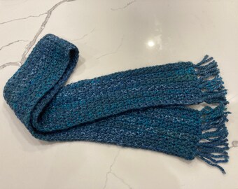 May include: A hand-knitted scarf in shades of blue and teal, with a textured appearance. The scarf has a fringed edge on both ends. The yarn has a speckled effect, adding visual interest to the accessory.