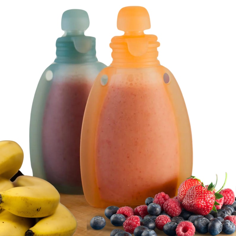May include: Two clear silicone squeeze bottles with orange and blue lids filled with pink and orange smoothies. The bottles have handles on the sides. The bottles are sitting on a wooden surface with a pile of yellow bananas and a scattering of blueberries, raspberries, and strawberries.
