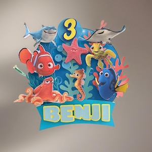 May include: A colorful cake topper featuring characters from the movie "Finding Nemo" and "Finding Dory". The design includes a blue background with the number 3, coral, and the name "BENJI".