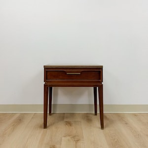 May include: A brown wooden nightstand featuring a single drawer with a horizontal metal pull handle. The piece stands on four straight legs and has a flat rectangular top surface, showcasing a smooth finish and clean, linear construction.