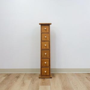 Könnte beinhalten: Ein hohes, schmales Aufbewahrungsmöbel aus Holz mit sechs übereinander angeordneten Schubladen. Jede Schublade ist aus hellbraunem Holz gefertigt und mit einem einzelnen, kleinen, runden, hellen Holzknopf in der Mitte der Frontblende versehen.