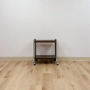 May include: A two-tier rolling trolley featuring a dark brown wood-grain finish on the shelves and a silver-toned metal frame. The trolley is supported by four small swivel castors attached to the base of the vertical metal supports on each side.
