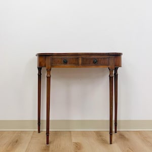 May include: A brown wooden console table with a semi-oval top and two drawers. The table features four slender, fluted legs and two dark circular drawer pulls. The wood grain is visible across the surface, and the design includes vertical grooved detailing on the front frame.