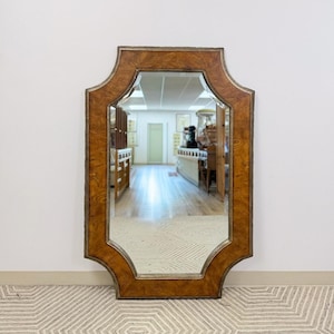 Puede incluir: Un espejo decorativo grande con un marco de forma única. El marco tiene una apariencia cálida de veta de madera con un borde dorado. El espejo es rectangular con una parte superior e inferior curvas. Está apoyado contra una pared blanca, reflejando una habitación.