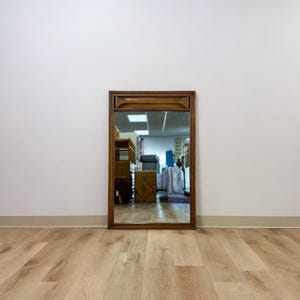 May include: A rectangular wall mirror with a medium-toned brown wooden frame. The top section of the frame features a recessed, horizontal decorative molding detail. The glass surface reflects an indoor space with various furniture items.