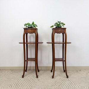 May include: Two antique wooden plant stands with three tiers each. The stands have a dark brown finish and ornate detailing on the top tier. Each stand holds a small potted plant with green leaves. The stands are placed against a white wall.