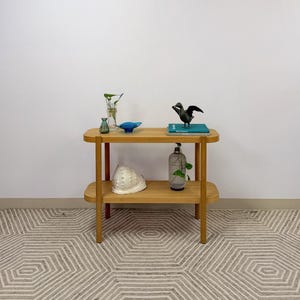 May include: A light wood two-tier console table with rounded edges. The top shelf holds a small clear glass vase with a plant, a blue glass bowl, and a dark bird figurine on a teal book. The bottom shelf holds a large white seashell and a glass vase.
