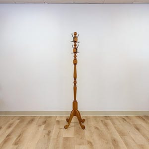 May include: A tall, free-standing wooden coat rack with a light brown finish. The structure features a central turned post, a tripod base with curved legs, and two tiers of metal hooks at the top for hanging garments and accessories.
