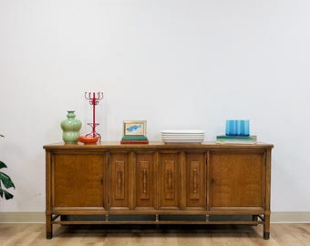 May include: A long wooden sideboard featuring four cabinet doors with vertical paneling and brass hardware. The top surface holds a light green vase, a red metal candle holder, a small framed fish print, white rectangular dishes, and blue glasses.