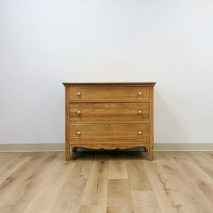 May include: A light brown wooden dresser featuring three horizontal drawers, each equipped with two small round knobs. The piece has a flat top surface and a decorative scalloped apron along the bottom edge, supported by four short, straight legs.