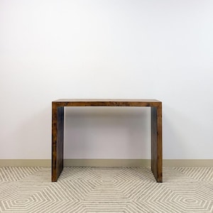 Può includere: Una consolle rettangolare in legno marrone scuro. Il tavolo ha un design semplice e moderno con quattro gambe e un piano piatto. Il tavolo è lungo circa 76 cm, largo 30 cm e alto 76 cm.