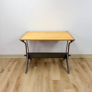 May include: A drafting table featuring a rectangular light-colored wooden top surface supported by a dark gray metal frame with angled legs and a horizontal crossbar, designed with adjustable hardware components on the sides.