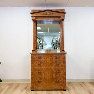 May include: A tall wooden dresser with a burl wood veneer finish, featuring four drawers with round metal pulls. The unit includes an integrated rectangular mirror framed by two columns and a decorative pediment with a central gold-toned emblem.