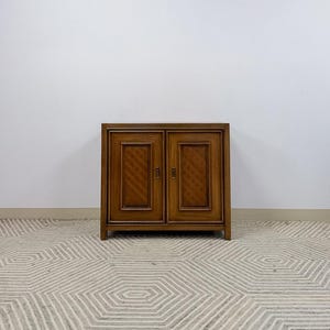 May include: A medium-brown wooden cabinet featuring two doors with recessed panels and a subtle diagonal grid pattern. The cabinet has a rectangular frame, clean lines, and small vertical metal handles centered on each door panel.