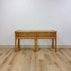 May include: A rectangular console table constructed from light brown natural rattan. The piece features a flat top surface and a decorative lattice-patterned apron supported by four vertical legs connected by a horizontal frame structure.