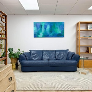 May include: A navy blue leather sofa with three cushions dominates the room. Above it hangs a large abstract painting in shades of blue and green. The room also features a patterned rug, wooden shelves, and a potted plant.