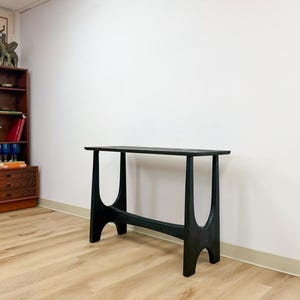 May include: A black wooden console table featuring a rectangular top surface supported by two U-shaped legs connected by a horizontal stretcher bar. The table has a visible wood grain texture and a dark finish, standing on a light-colored floor.
