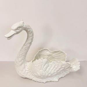 Peut inclure: Un bol en céramique blanche en forme de cygne présentant des textures de plumes moulées détaillées sur le cou, le corps et les ailes. Le cygne a un cou arqué, un bec ouvert et un centre creux conçu pour contenir des objets, avec une finition lisse.