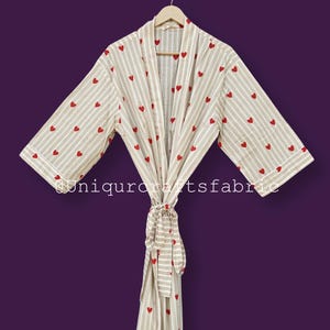 May include: A beige and white striped robe with red heart accents. The robe is hanging on a wooden hanger against a purple background. The robe has a matching tie belt.