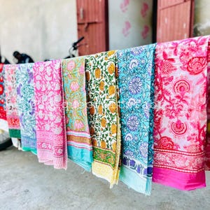 Pu&ograve; includere: Una collezione di sei sciarpe di cotone colorate con motivi floreali appese a uno stendino. Le sciarpe sono in tonalit&agrave; di rosa, blu, verde e giallo.