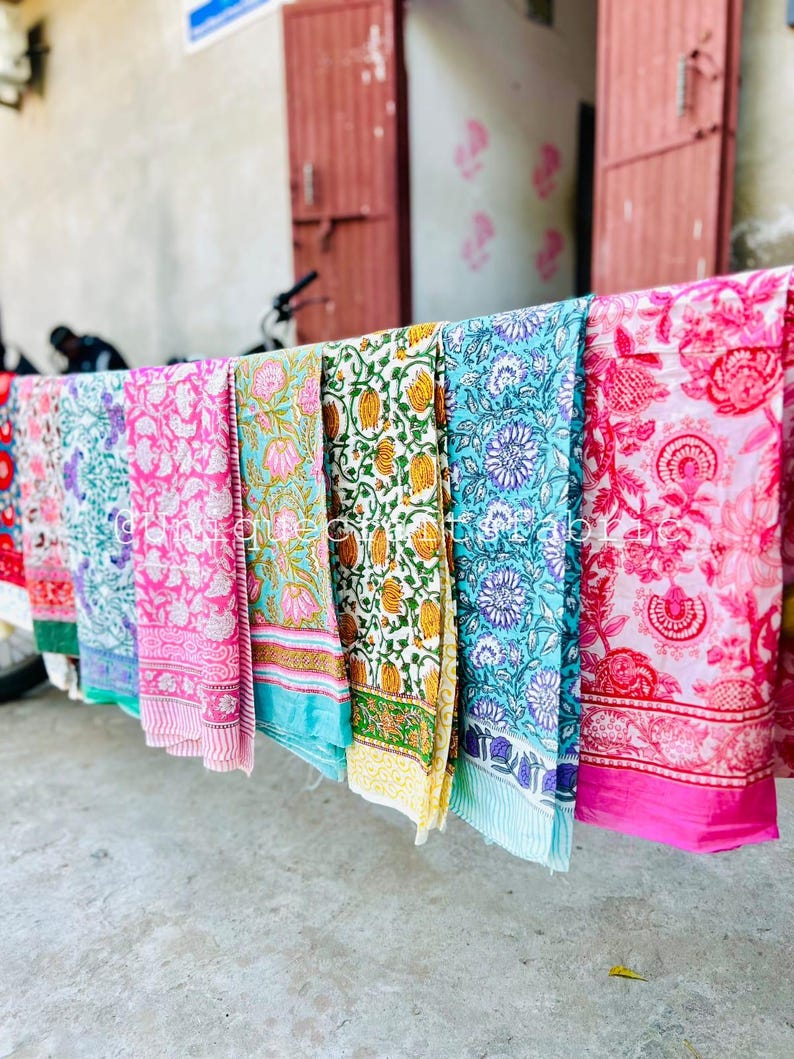 Pu&ograve; includere: Una collezione di sei sciarpe di cotone colorate con motivi floreali appese a uno stendino. Le sciarpe sono in tonalit&agrave; di rosa, blu, verde e giallo.
