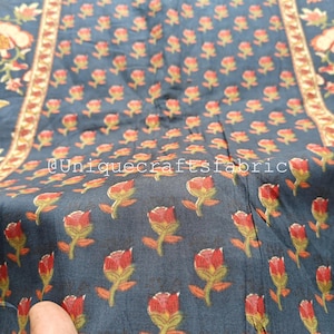 Op de afbeelding: Een close-up van een donkerblauwe stof met een herhalend patroon van kleine rode bloemen. De stof heeft een subtiele glans en een zachte textuur. De tekst "@uniquecraftsfabric" is zichtbaar in de linkerbenedenhoek van de afbeelding.