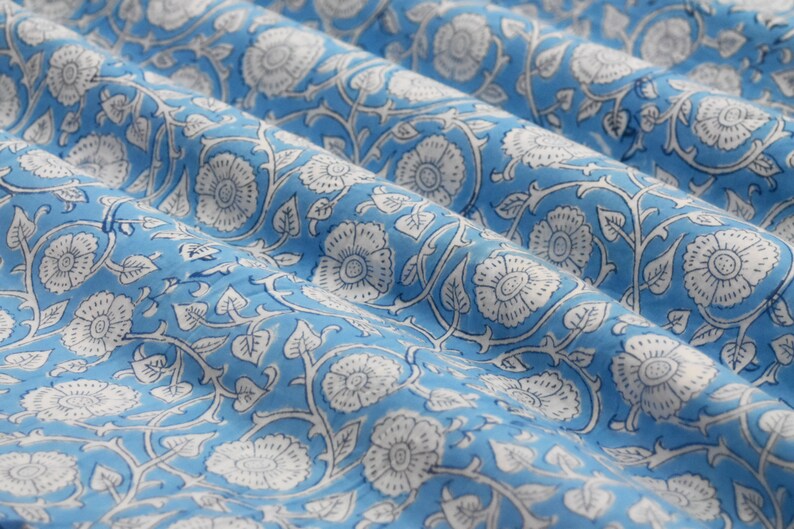 May include: Blue fabric with a white floral pattern. The fabric has a repeating pattern of white flowers and leaves on a light blue background.