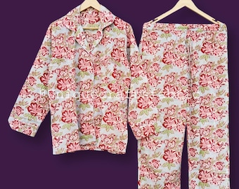 Könnte beinhalten: Ein zweiteiliger Schlafanzug mit floralem Muster. Das Set besteht aus einem Hemd mit Knöpfen und einer passenden Hose, beide in Hellgrau mit einem sich wiederholenden Muster aus roten und grünen Blumen. Die Pyjamas hängen an Holzbügeln vor einem violetten Hintergrund.